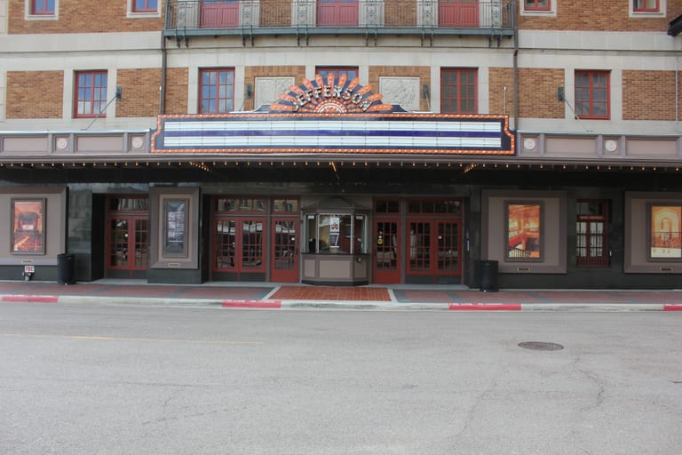 The 1927 Jefferson Theatre facade in downtown Beaumont, with its illuminated sunburst marquee reading JEFFERSON above red-trimmed doors and a street-level box office kiosk.
