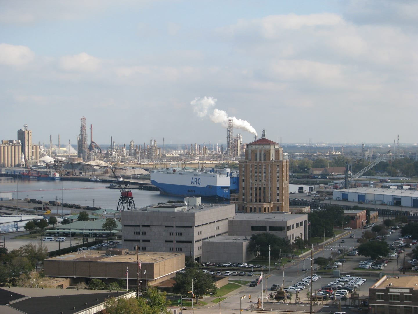 Elevated view of the Port of Beaumont: a blue and white roll-on/roll-off car carrier docked on the Neches River, refineries and a steam plume behind it, and the tan Jefferson County courthouse tower rising above downtown.